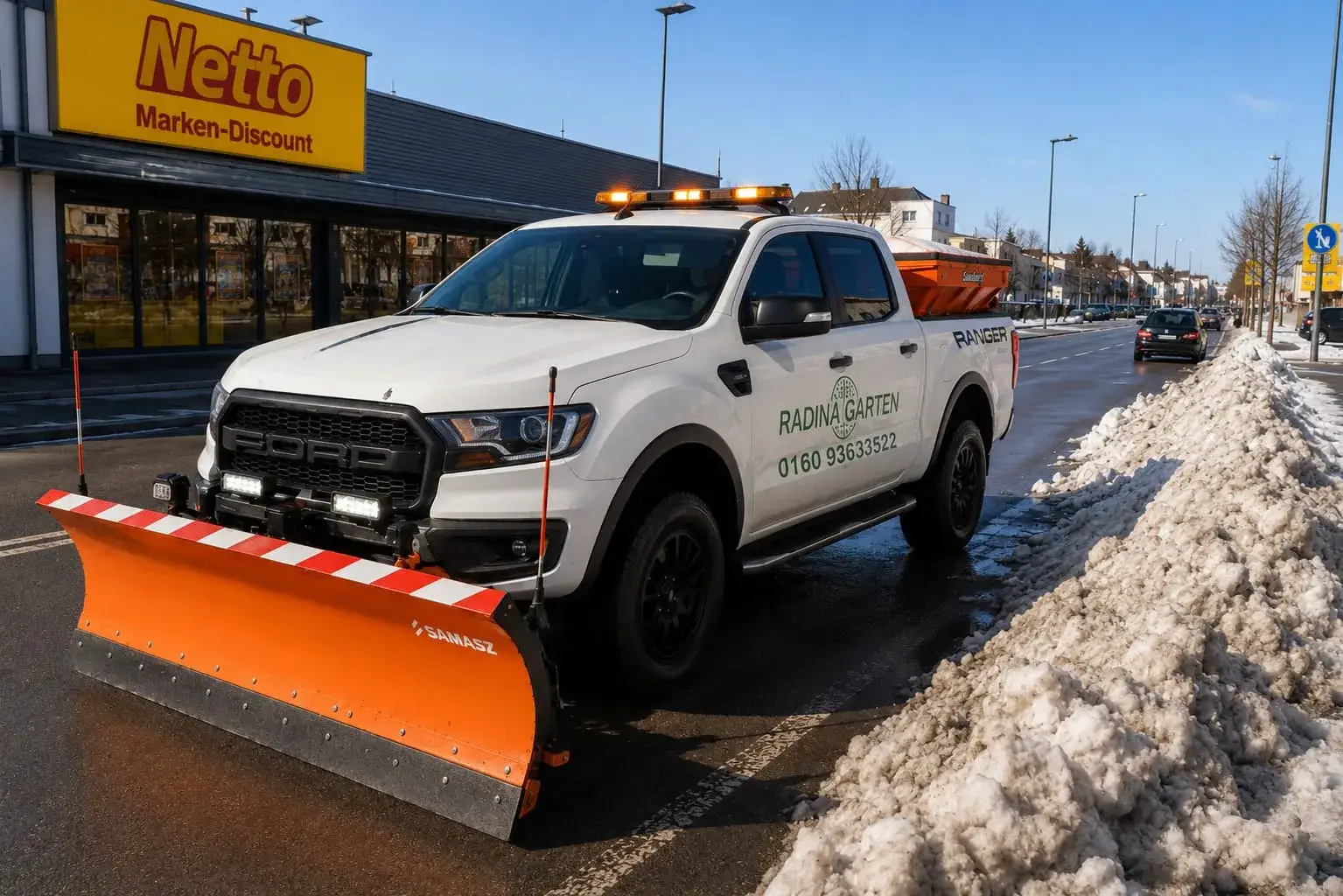 Winterdienst-Pickup mit Streuer neben geräumtem Schneehaufen am Supermarkt